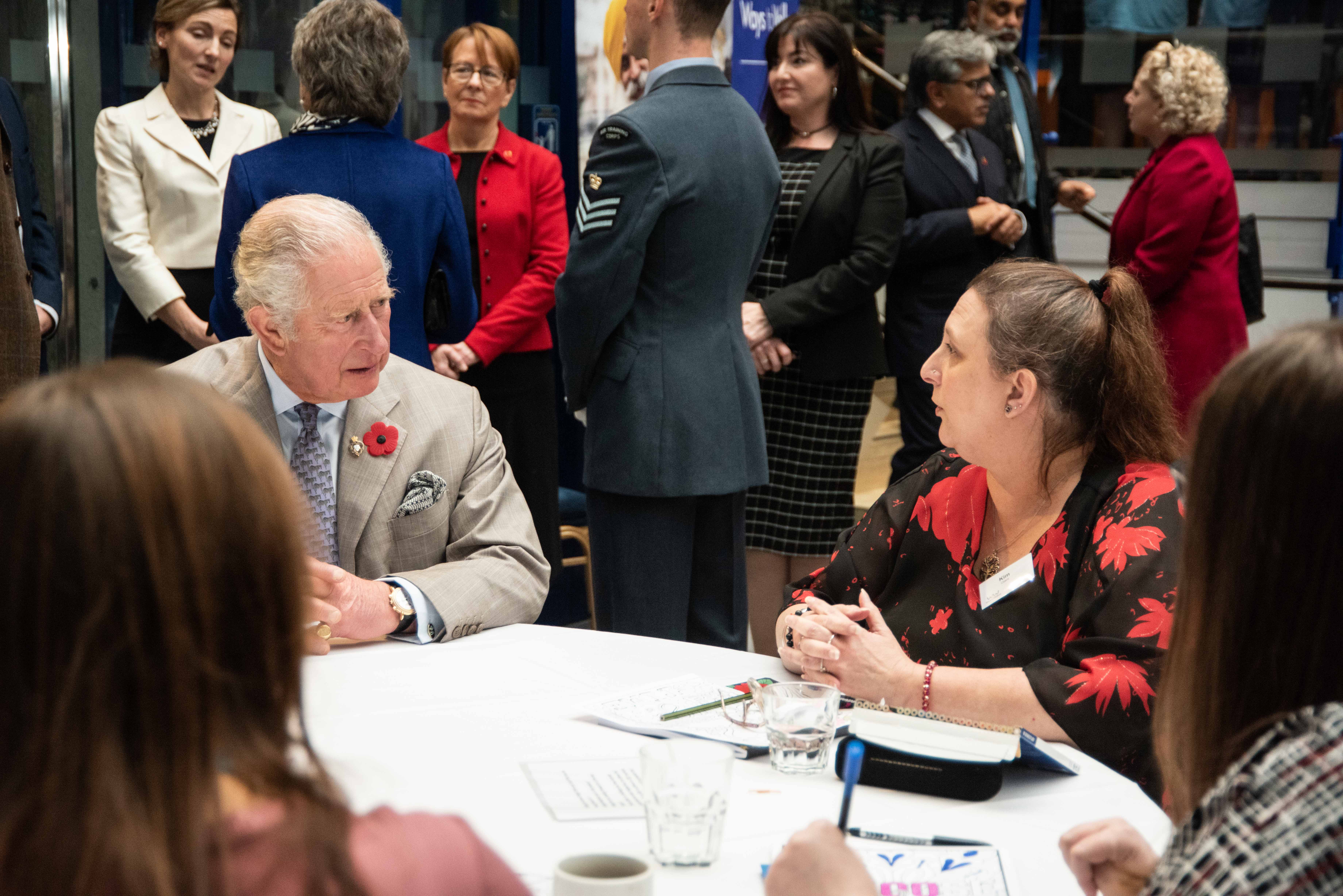 Prince Charles sits at table speaking to lady in red and black blouse