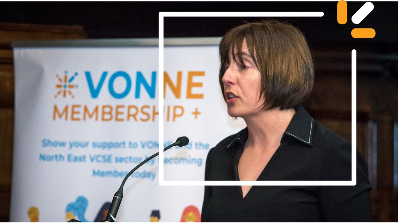 Photo of Alison speaking into a microphone at VONNEs conference, with a VONNE membership banner in the background