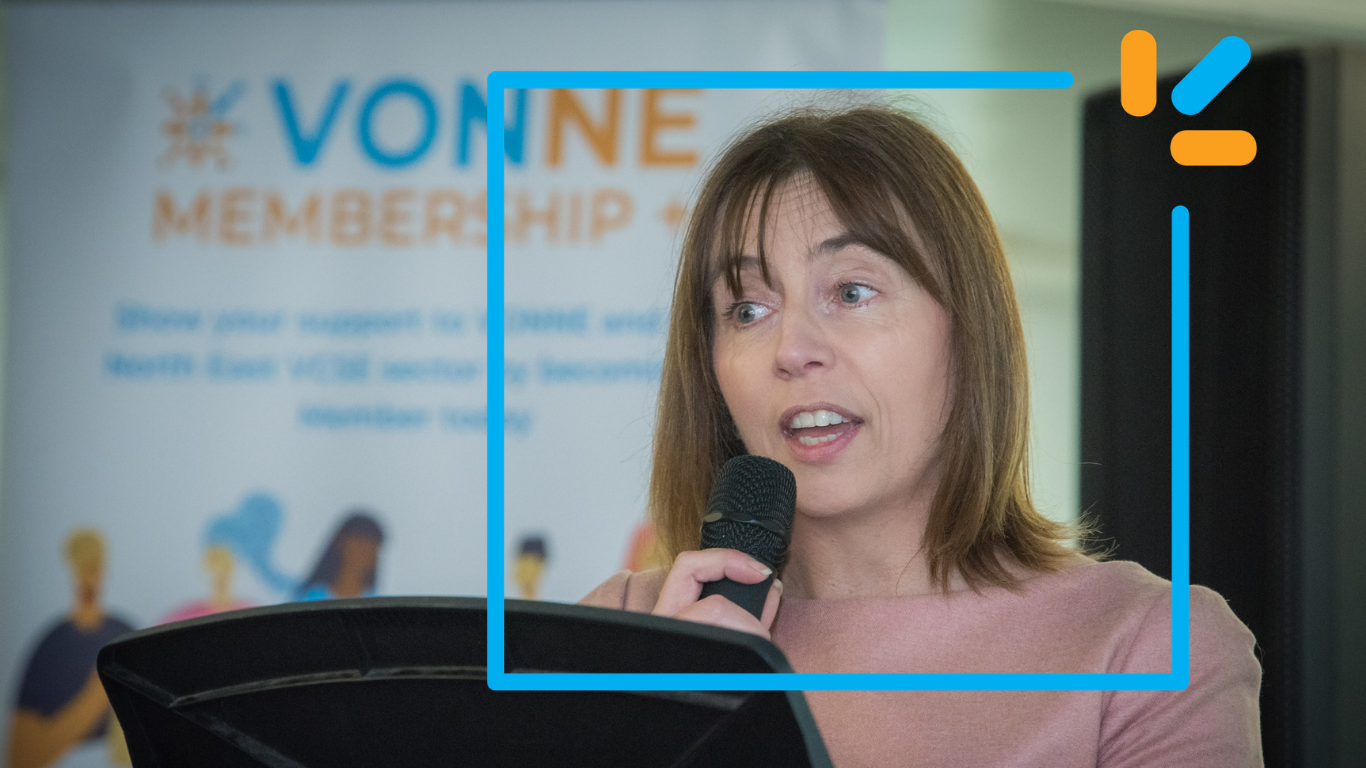 Photo of Alison speaking into a microphone at VONNEs conference, with a VONNE membership banner in the background