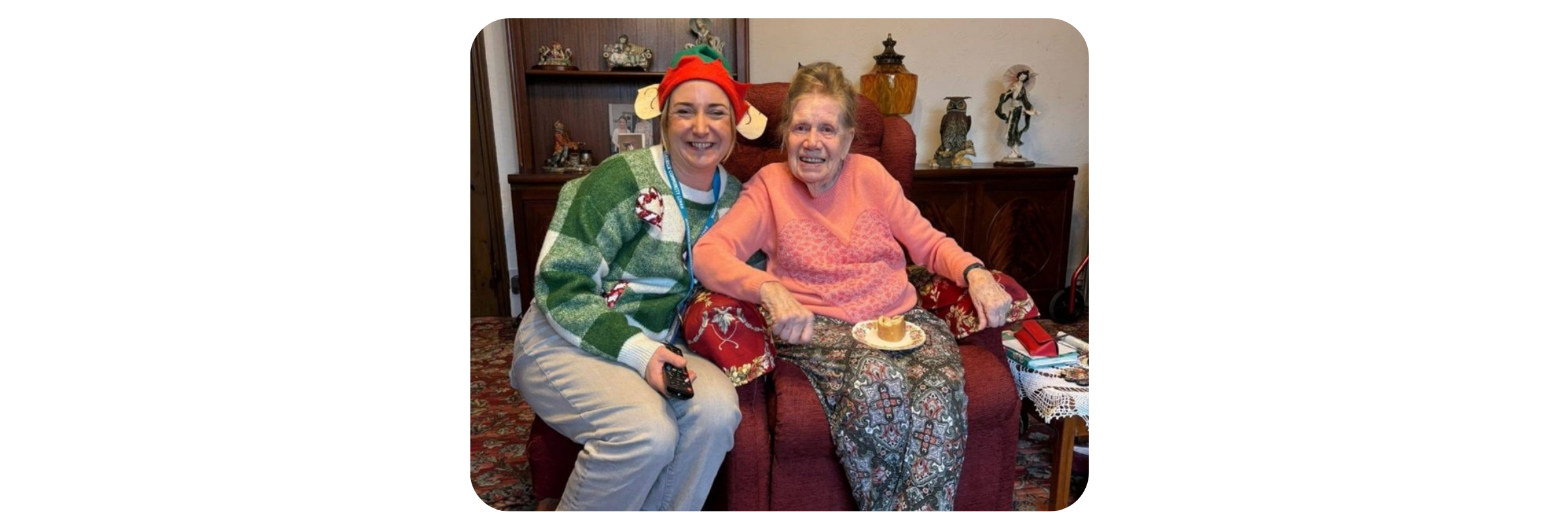 An older lady sitting in a comfy chair at home with a patterned carpet and lots of photos and ornaments on a sideboard. She's eating a mince pie and kneeling next to her chair holding a christmas gift is a woman dressed in a christmas elf jumper and a hat with elf ears. 
